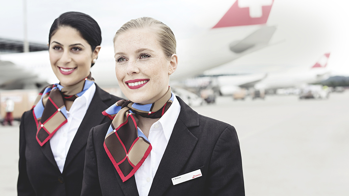 Two smiling SWISS cabin crew members on a runway, behind them several SWISS aircraft