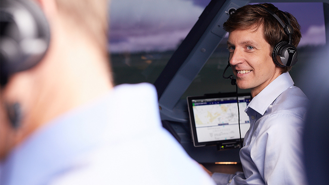 A man wearing headphones sits in an aircraft cockpit and smilingly looks at his colleague