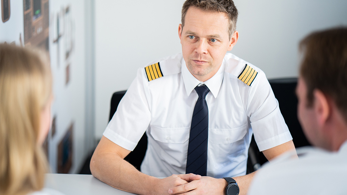 A pilot sits at a table, where he has a conversation with a woman and a man