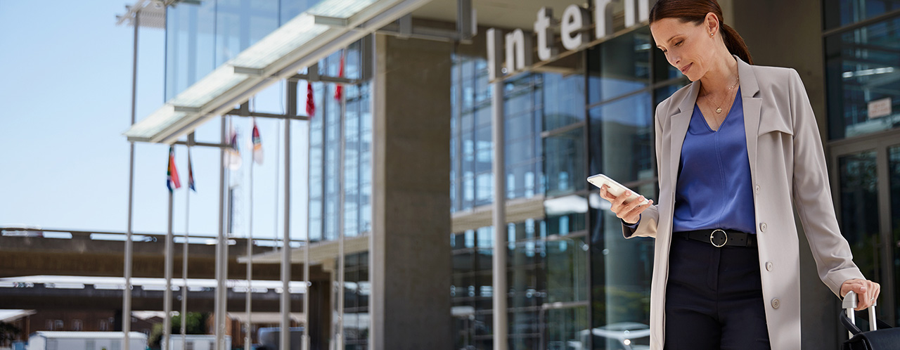 A businesswoman stands with her suitcase on wheels in front of an airport entrance and looks at her smartphone