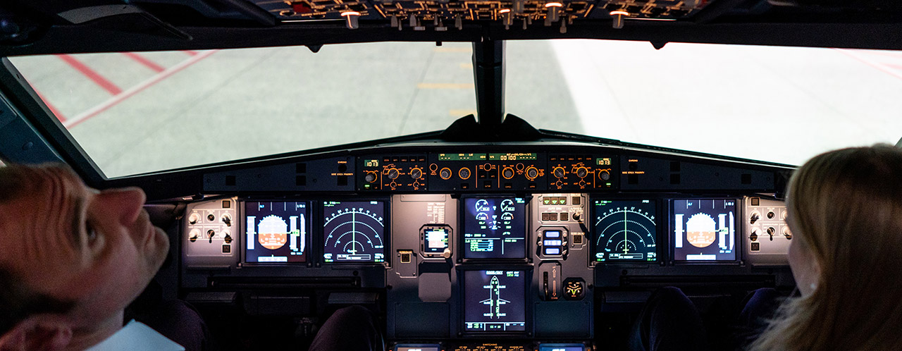 Two people sit in an aircraft cockpit, one person looks up to the control instruments