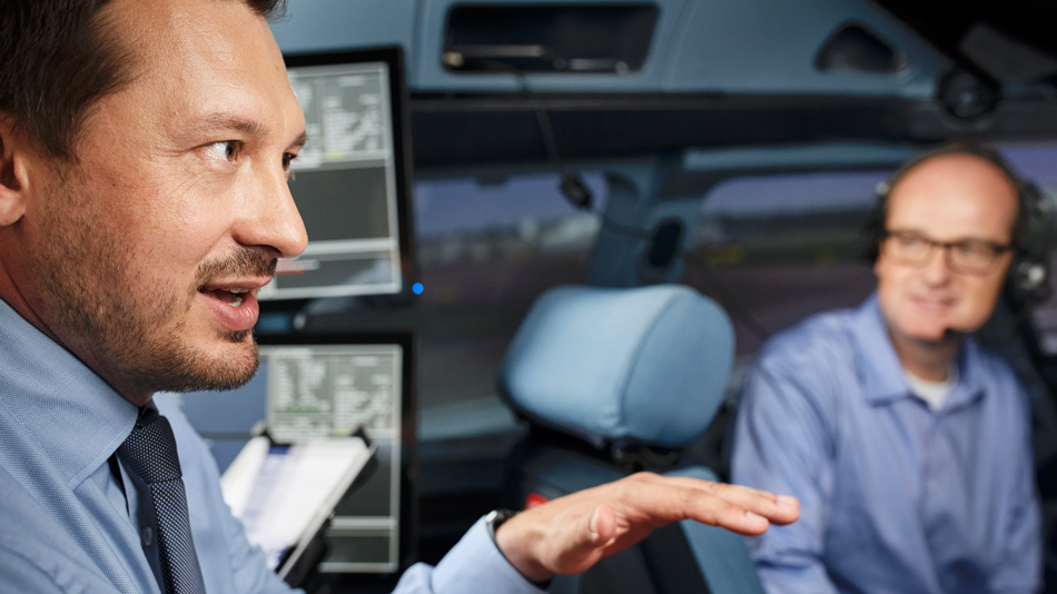 Two men sit in a cockpit, one of them explains something