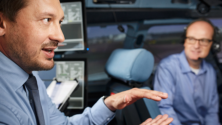A view into the cockpit of a full flight simulator, where a pilot is seated and an instructor is providing an explanation.