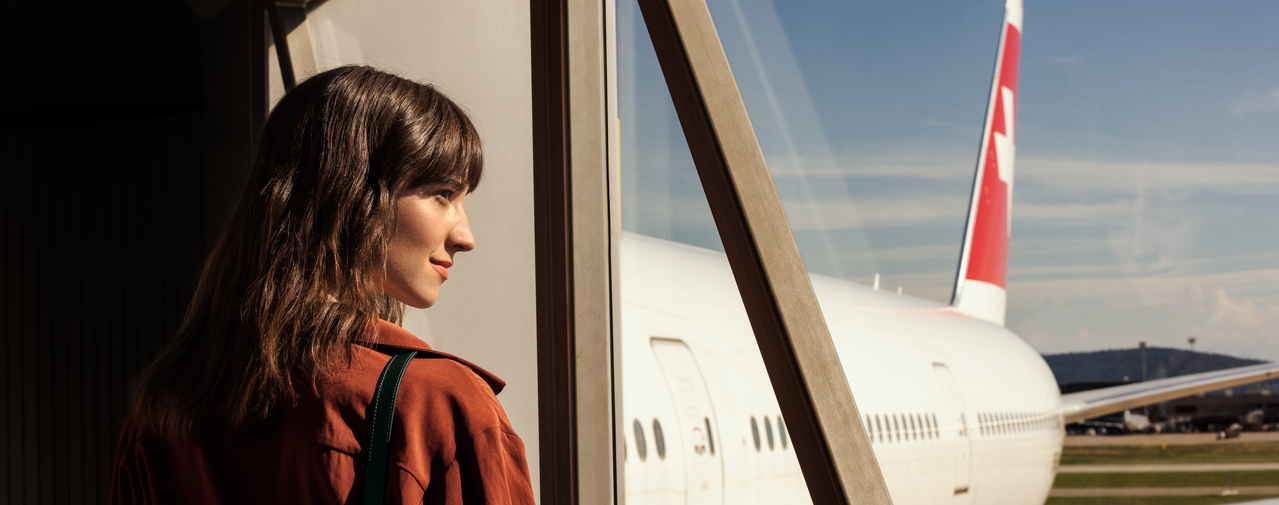 Eine Person steht an einem Flughafen und blickt aus dem Fenster, vor dem Fenster steht ein Flugzeug mit SWISS Branding.