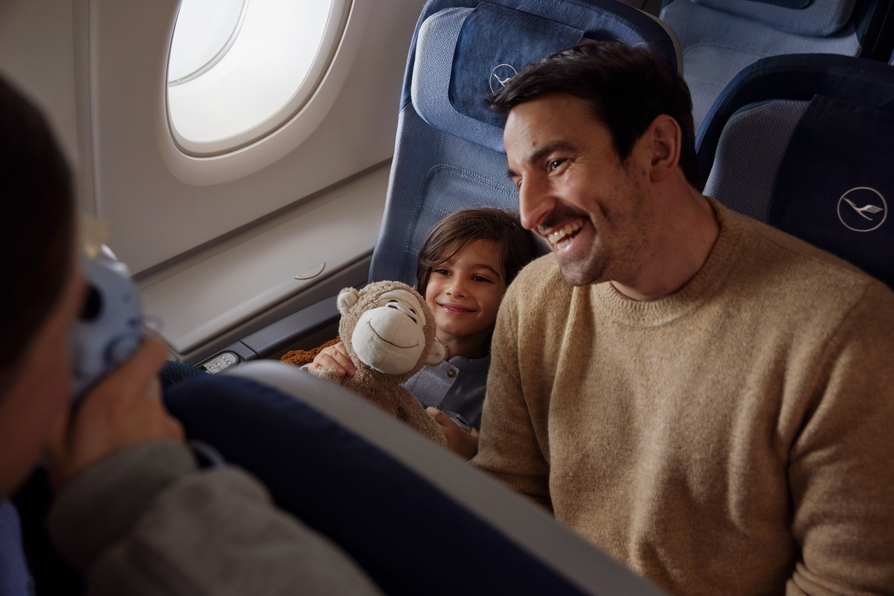 Ein Vater sitzt neben seinem Kind im Flugzeug, vom Vordersitz fotografiert eine Person die beiden.