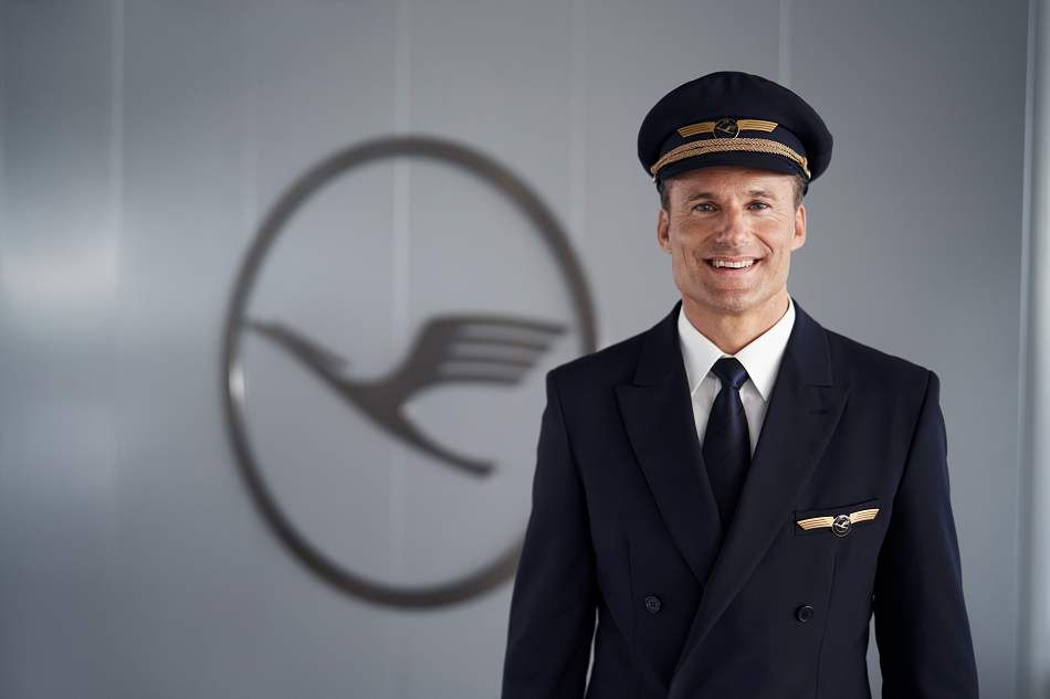 Ein Pilot steht vor dem Kranich Logo der Lufthansa und lächelt in die Kamera.