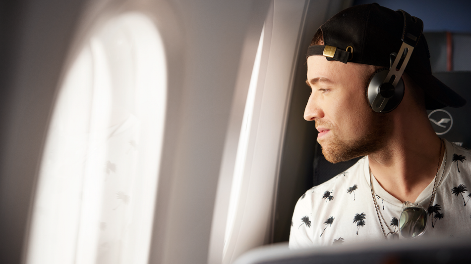 Eine Person mit Kopfhörern und Cap sitzt in einem Flugzeug am Fenster und blickt aus dem Fenster.