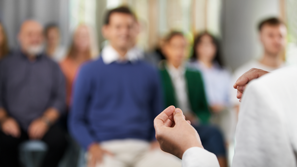 Im Hintergrund sitzen Personen im Publikum, im Vordergrund die Hände und der Rücken einer Person, die zum Publikum spricht.