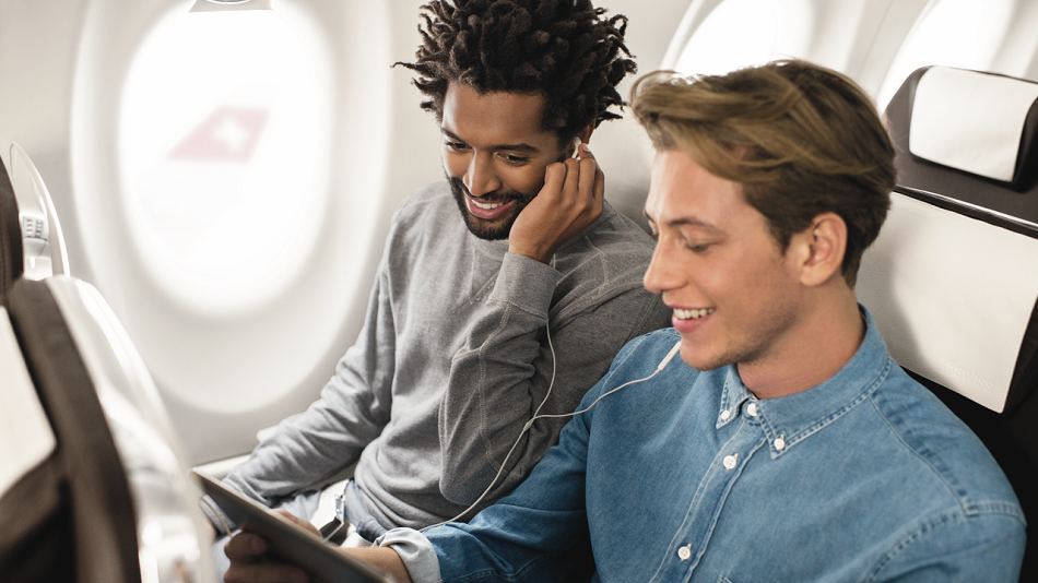 Zwei Personen sitzen gut gelaunt nebeneinander im Flugzeug, teilen sich einen Kopfhörer und schauen auf ein Tablet.